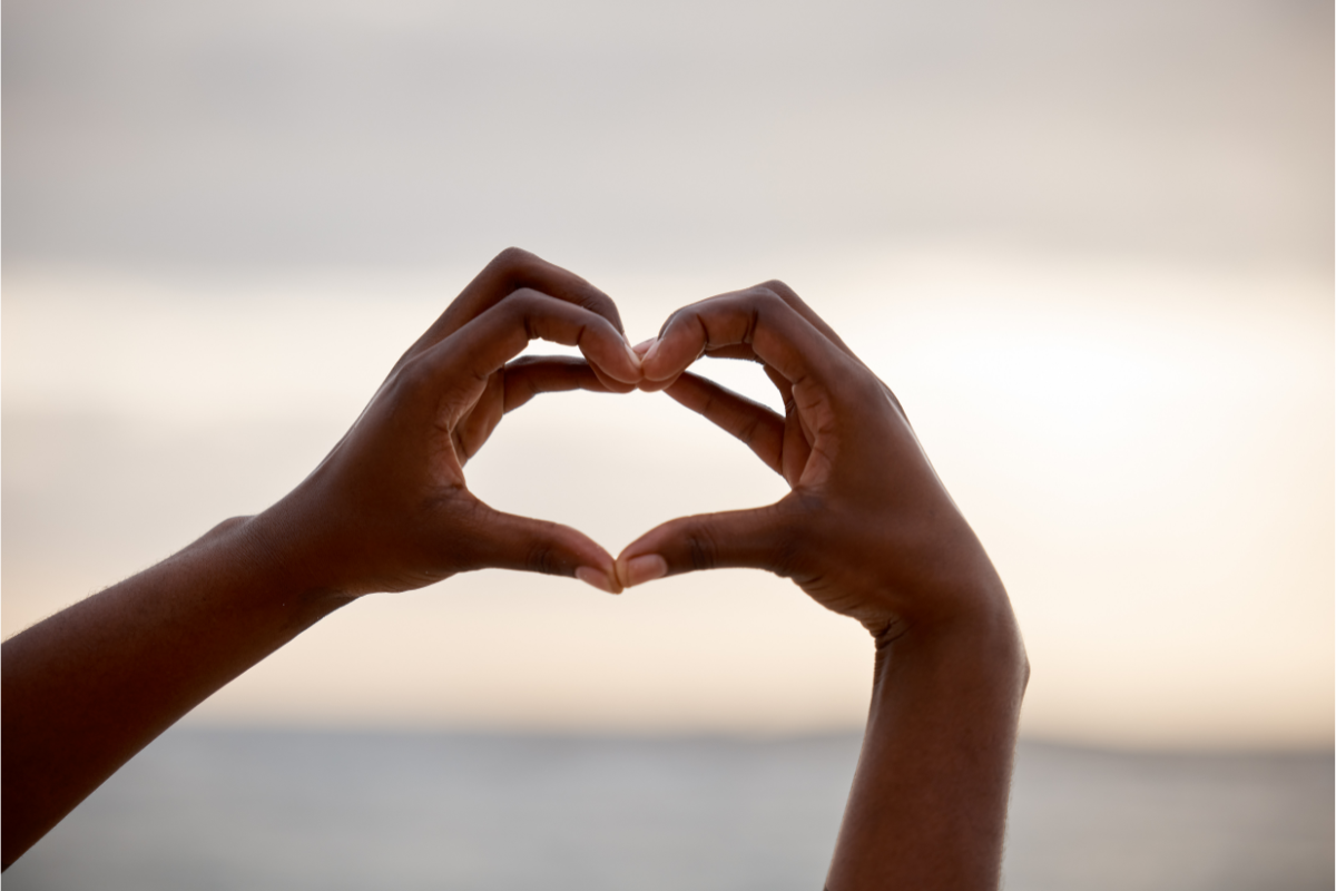 Hands forming a heart shape against a blurred ocean and sky background, symbolizing love, connection, and positivity.
