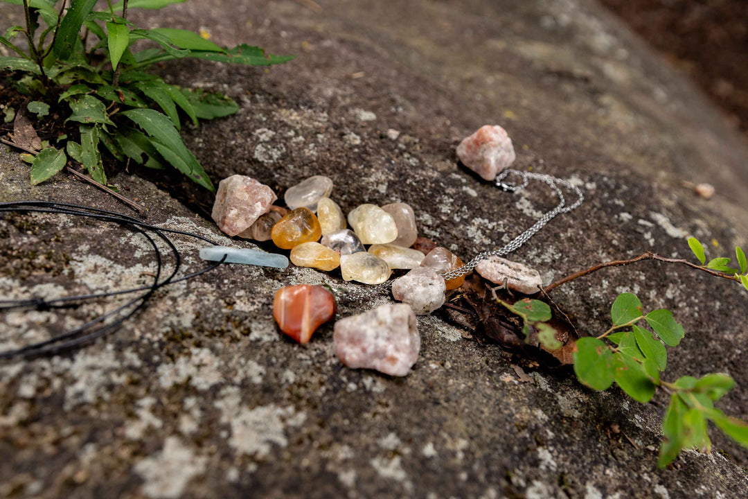 Assorted tumbled and raw crystals including citrine, sunstone, and carnelian displayed on a natural rock surface outdoors with crystal pendants on black cord and silver chain necklaces.