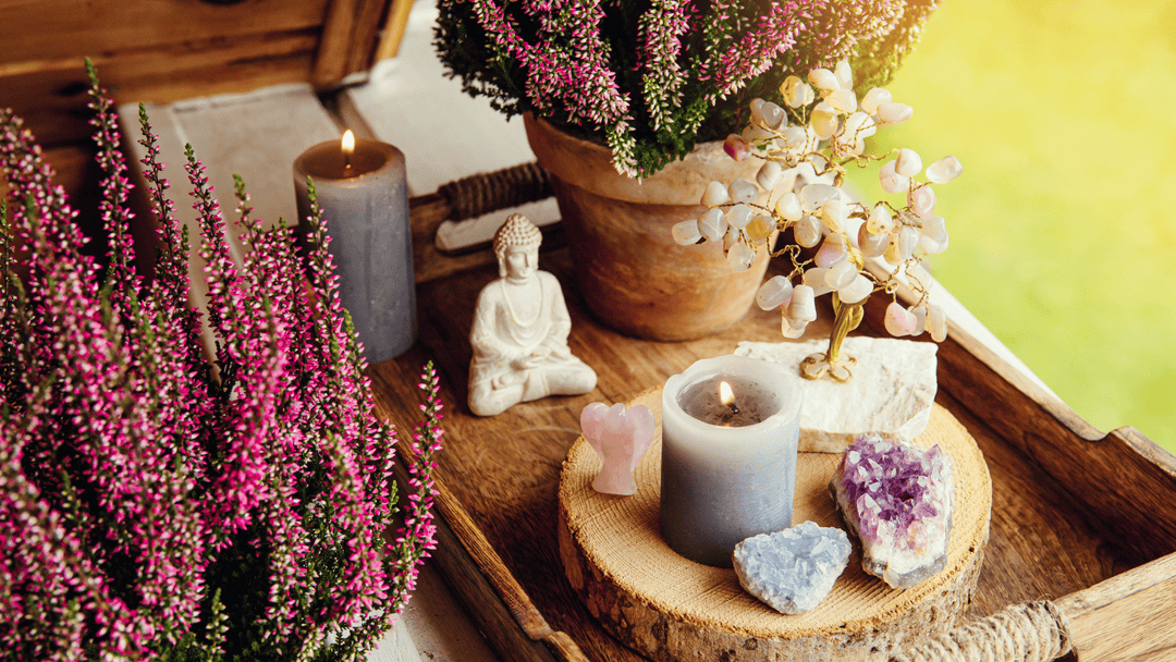Healing crystals, candles, and a Buddha statue arranged on a wooden tray beside pink heather flowers, creating a peaceful home altar by the window.