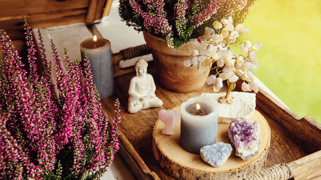 Healing crystals, candles, and a Buddha statue arranged on a wooden tray beside pink heather flowers, creating a peaceful home altar by the window.