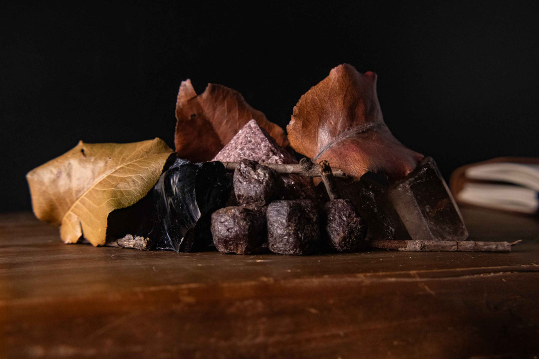 A dark moody flat-lay display featuring dried autumn leaves, dark obsidian, raw garnets, a lepidolite pyramid, and smoky quartz crystal on a rustic wooden surface.