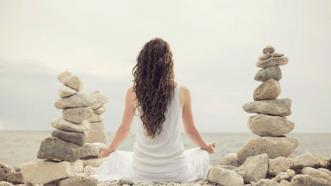 Woman meditating by the sea surrounded by balanced stone stacks, symbolizing peace, grounding, and good energy