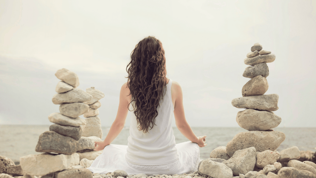 Woman meditating by the sea surrounded by balanced stone stacks, symbolizing peace, grounding, and good energy