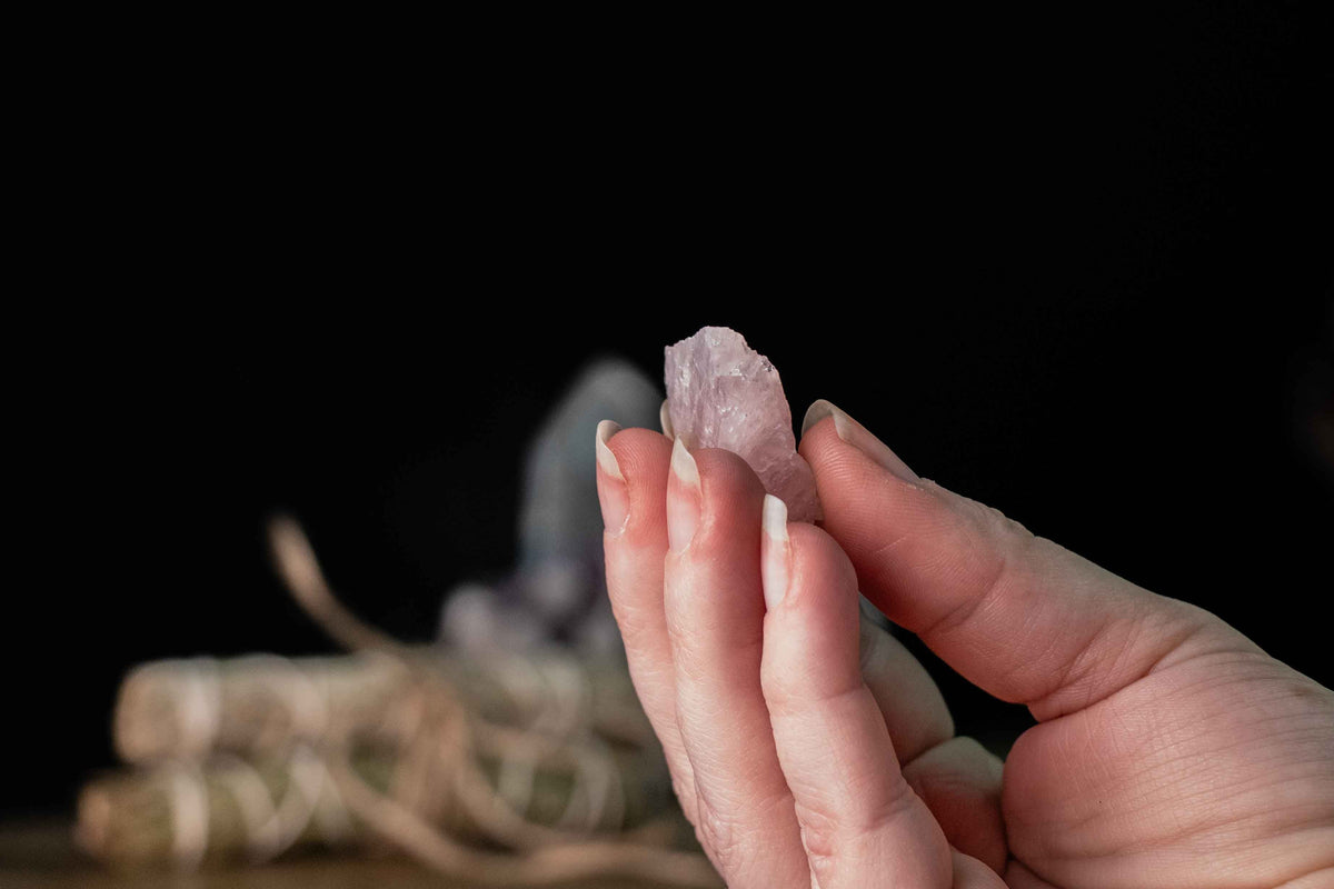 Hand holding a small raw pink crystal against a dark background, with a blurred sage bundle resting on a wooden surface behind it, suggesting a crystal cleansing ritual.