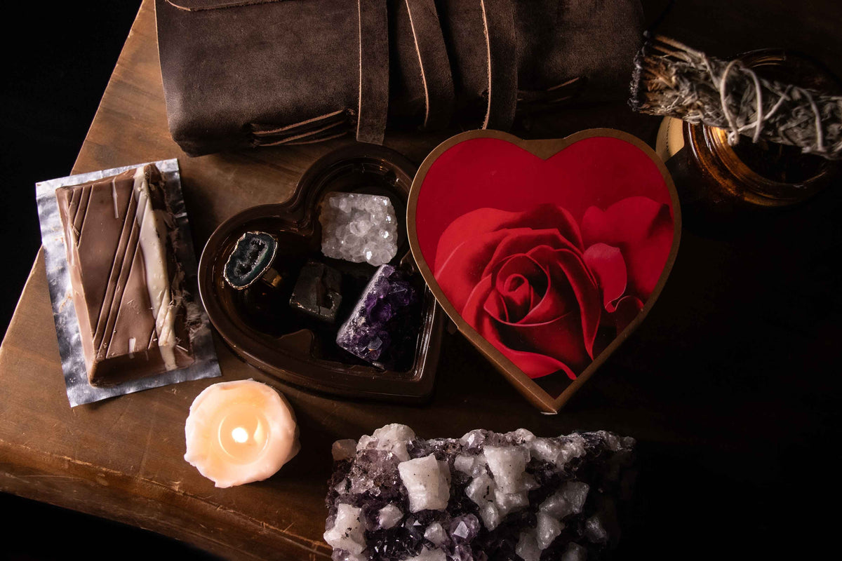 Ritual setup on a wooden table featuring a heart-shaped box, crystals including amethyst and clear quartz, sage bundles, a lit white candle, a leather journal, and soft smoke rising in a moody setting.