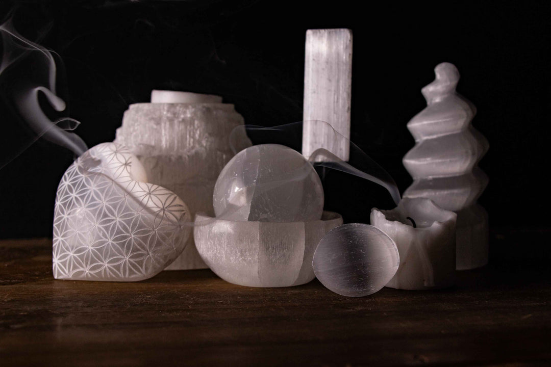 A collection of white Selenite crystals, including a Flower of Life engraved heart, a sphere in a bowl, and a tower, set against a dark background with ethereal smoke curls.