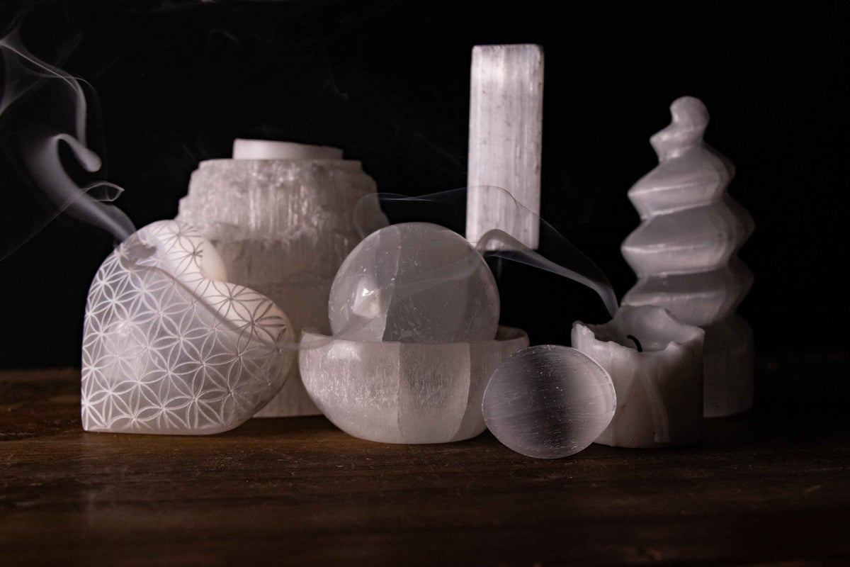 A collection of white Selenite crystals, including a Flower of Life engraved heart, a sphere in a bowl, and a tower, set against a dark background with ethereal smoke curls.