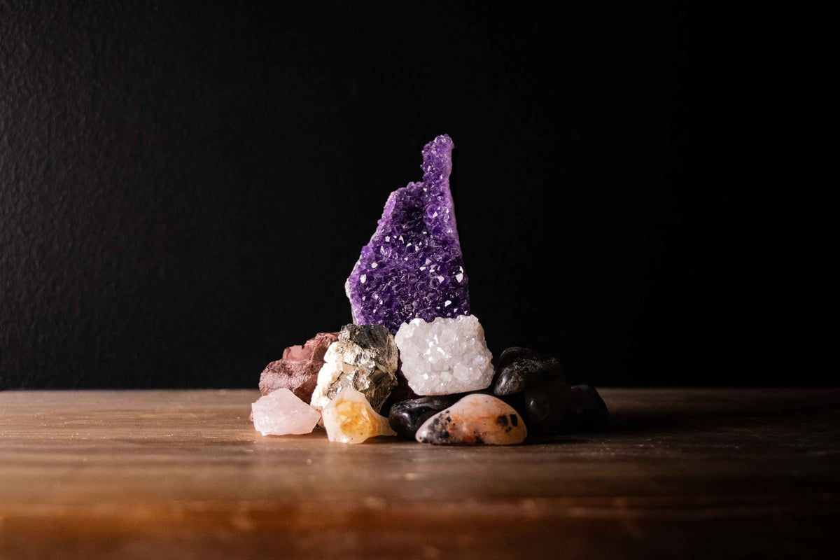 A stacked collection of various crystal forms including a tall, rough purple amethyst cluster, a sparkling white geode piece, shiny metallic pyrite, and smooth, polished tumbled stones on a wooden surface against a dark background.