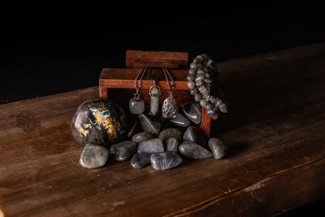 Labradorite crystal collection styled on dark wood surface featuring tumbled stones, sphere with gold and blue flash, pendants, chip bracelet, and ring displayed with wooden box against black background.