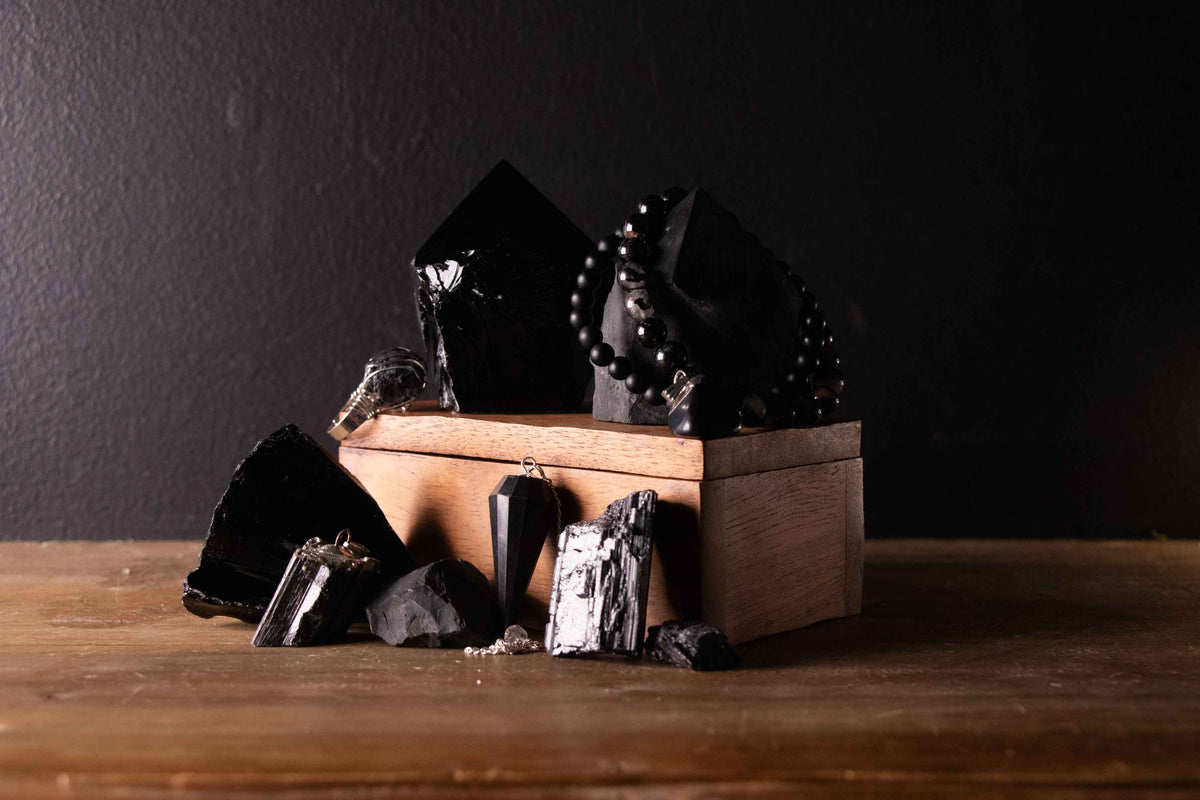 Black crystal collection flat lay on a wooden surface featuring obsidian points, raw black tourmaline, shungite pieces, a black obsidian pendulum and a black bead bracelet styled around a wooden box against a dark background.