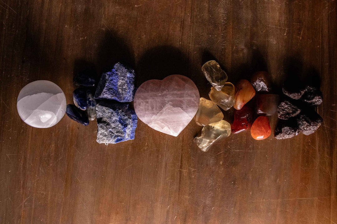 Row of seven chakra crystals arranged in a color gradient from crown to root, featuring selenite, sodalite, lapis, rose quartz, citrine, carnelian, and garnet on a dark wood surface.