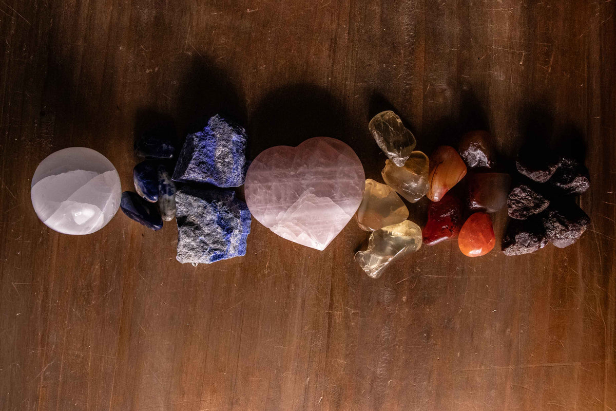 Row of seven chakra crystals arranged in a color gradient from crown to root, featuring selenite, sodalite, lapis, rose quartz, citrine, carnelian, and garnet on a dark wood surface.