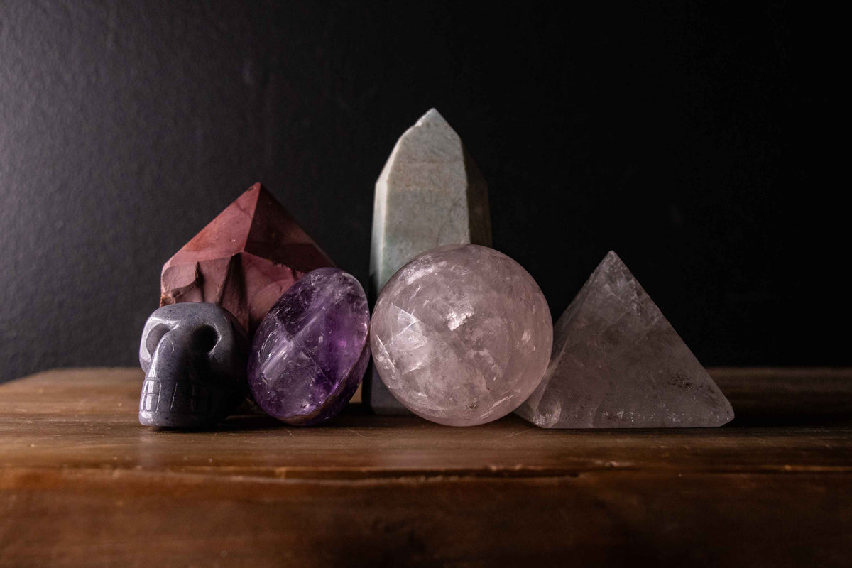 A collection of polished and shaped crystals arranged on a wooden surface against a dark background, including a small skull, an amethyst stone, a quartz sphere, a pyramid, and two tall points.