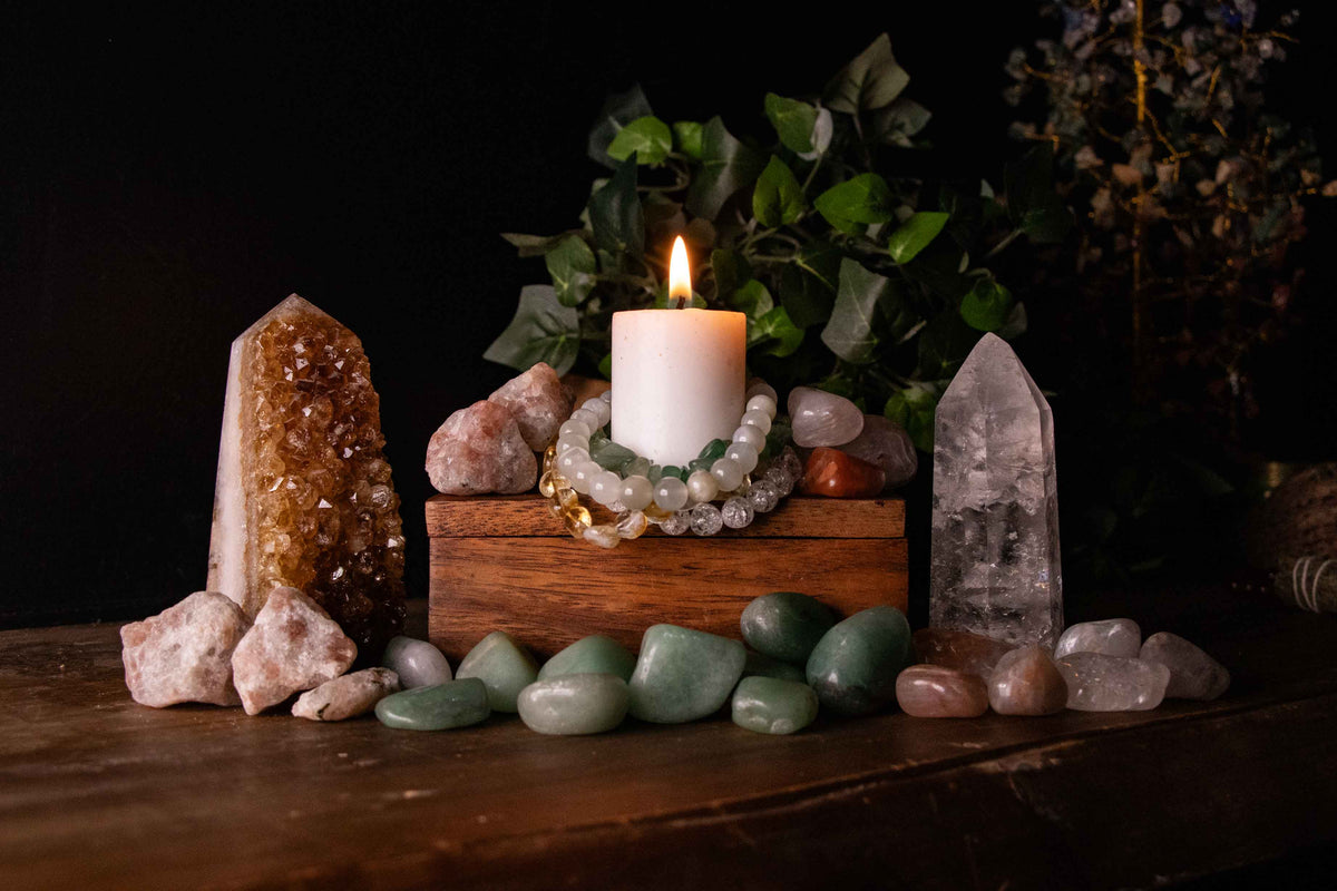 Wide shot of a lit candle surrounded by citrine cluster, clear quartz point, sunstone, moonstone and green aventurine bracelet, and tumbled crystals on a wooden box, styled with greenery.