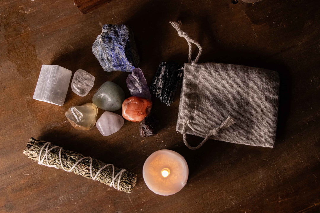 Overhead flat lay of selenite, clear quartz, lapis, amethyst, carnelian, black tourmaline, sage, a candle, and a linen pouch arranged on a wooden table.