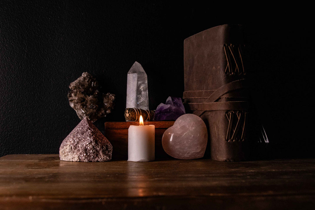 Moody dark altar flatlay featuring a smoky quartz crystal cluster, natural pyrite cluster, clear quartz point, white sage bundle, raw amethyst cluster, lit white pillar candle, polished rose quartz heart, and a leather bound journal, arranged on a dark wo