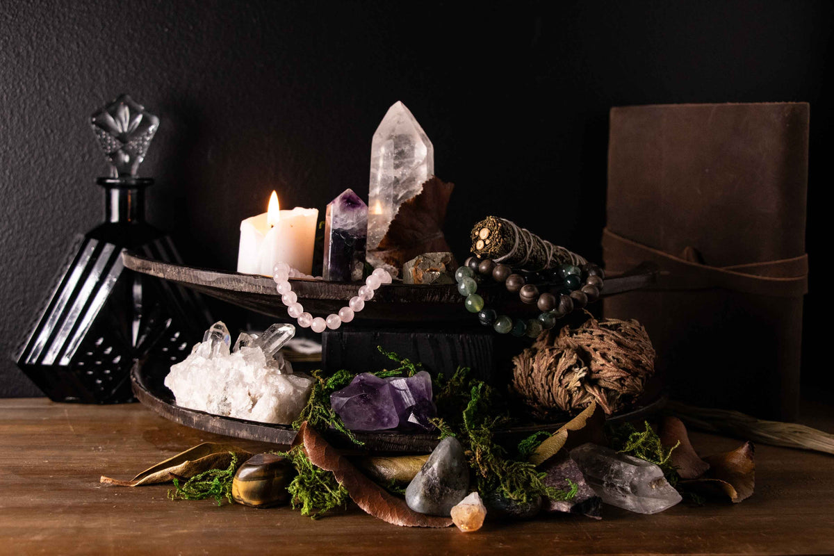 Crystal altar arrangement with a clear quartz point, amethyst point, raw amethyst cluster, clear quartz cluster, rose quartz bracelet, moss agate and petrified wood bracelet, sage bundles, moss, dried leaves, and a lit candle on a dark tiered stand.