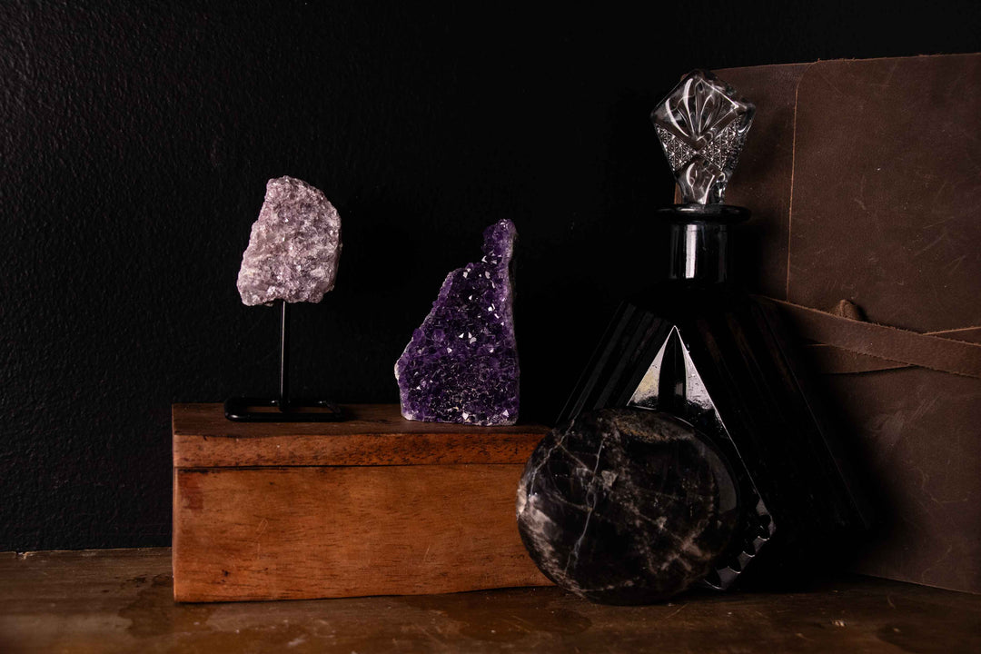 Lepidolite on a display stand, an amethyst crystal cluster, and a black moonstone sphere arranged on a wooden box, with a decorative black glass bottle in the background against a black backdrop.