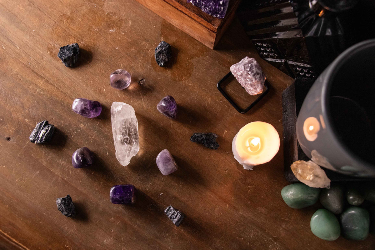 A crystal grid layout set on a warm wooden surface featuring a clear quartz point at the center, surrounded by an oval arrangement of polished purple amethyst stones and an outer boundary of rough black tourmaline pieces. To the right, a lit white candle 