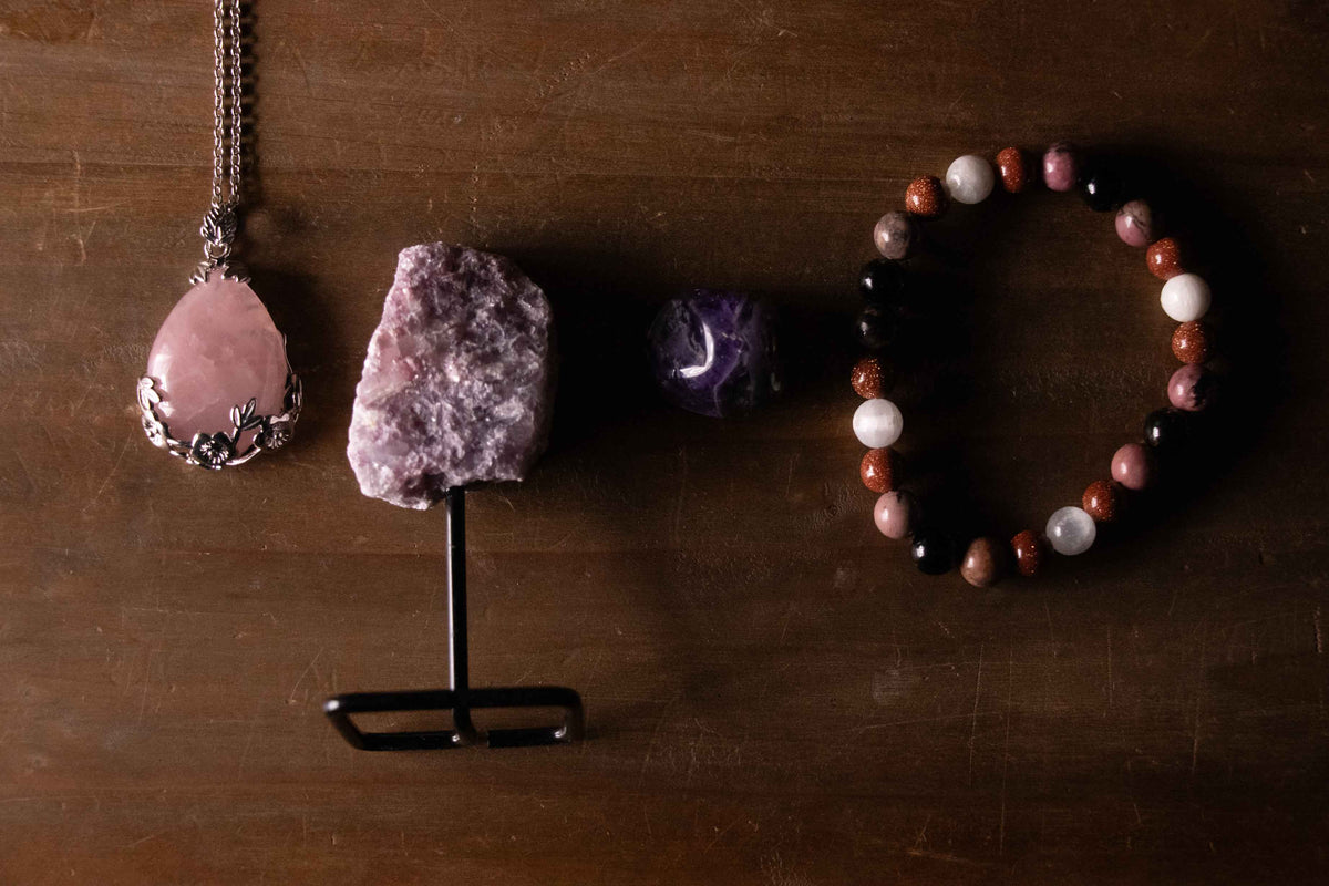 Flat lay of rose quartz pendant necklace, raw crystal on a stand, small amethyst stone, and crystal bead bracelet arranged on a wooden surface in soft, moody lighting.