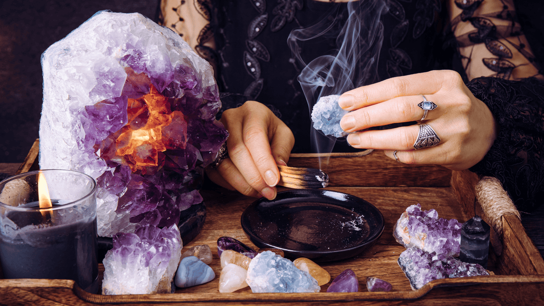 Woman cleansing crystals with Palo Santo smoke beside a glowing Amethyst geode, candle, and assorted healing stones on a wooden tray.