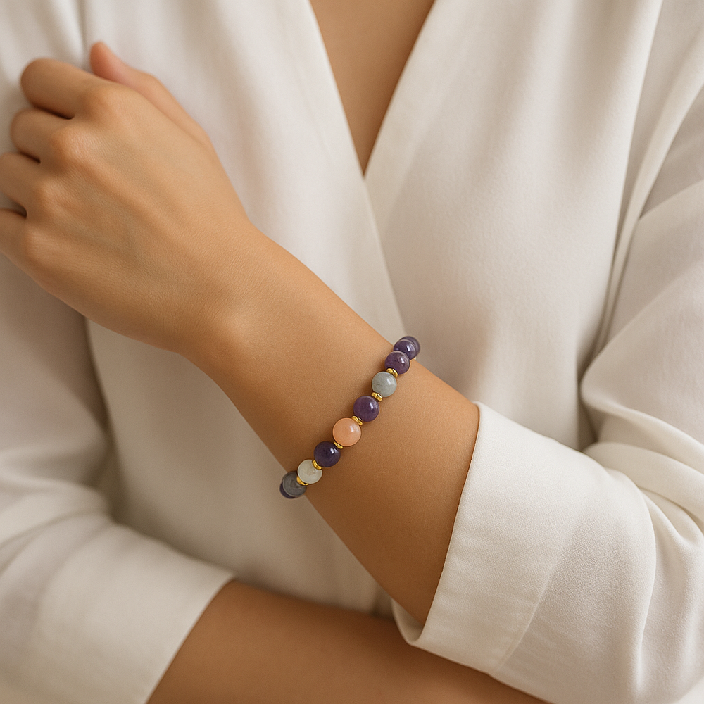 Close-up shot of a woman's arm wearing a stretch beaded bracelet featuring polished round amethyst, aquamarine, and peach moonstone beads interspersed with small gold spacers, against a crisp white shirt