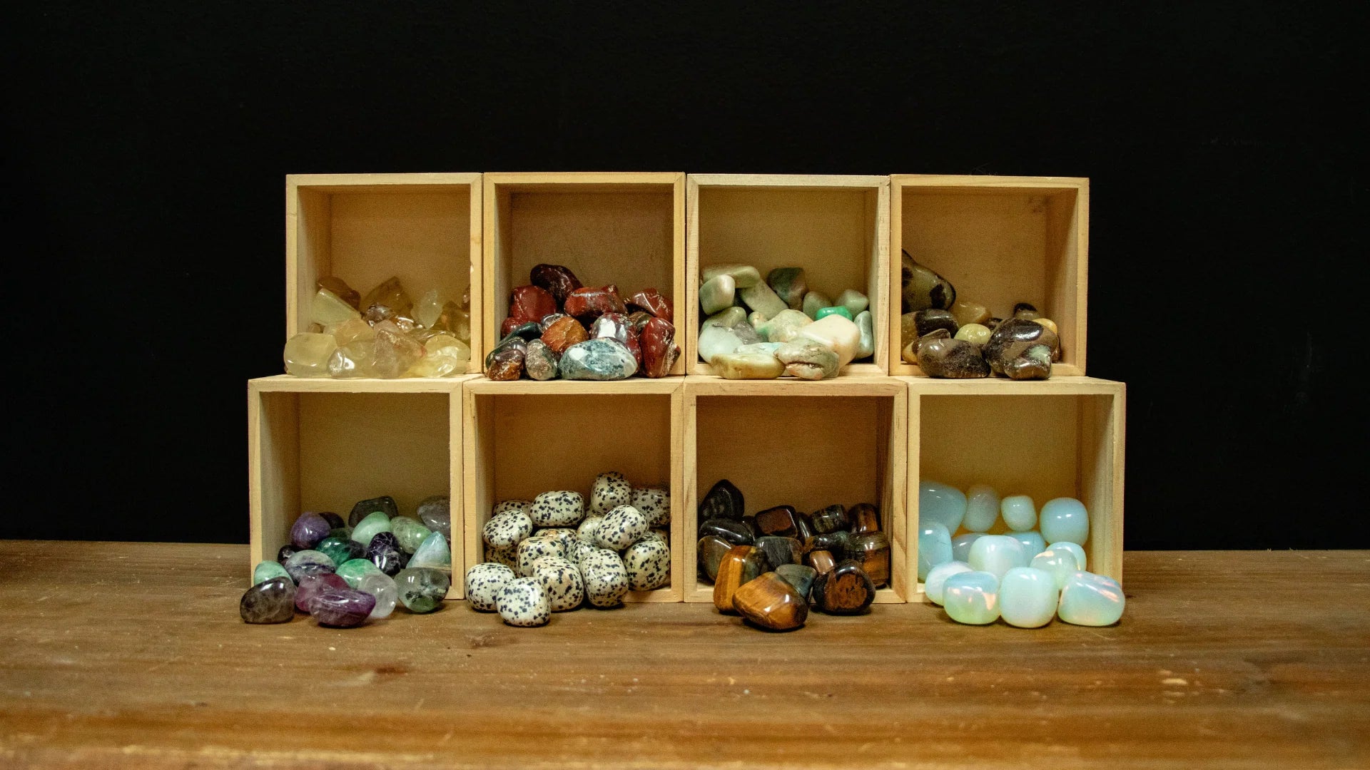 An organized display of eight small, light-wood square cubbies stacked in two rows on a rustic wooden tabletop against a solid black background. Each cubby is filled with a different variety of polished tumbled stones, including golden citrine, red jasper, light green aventurine, dalmatian jasper, tiger's eye, and glowing opalite, spilling slightly forward toward the viewer.