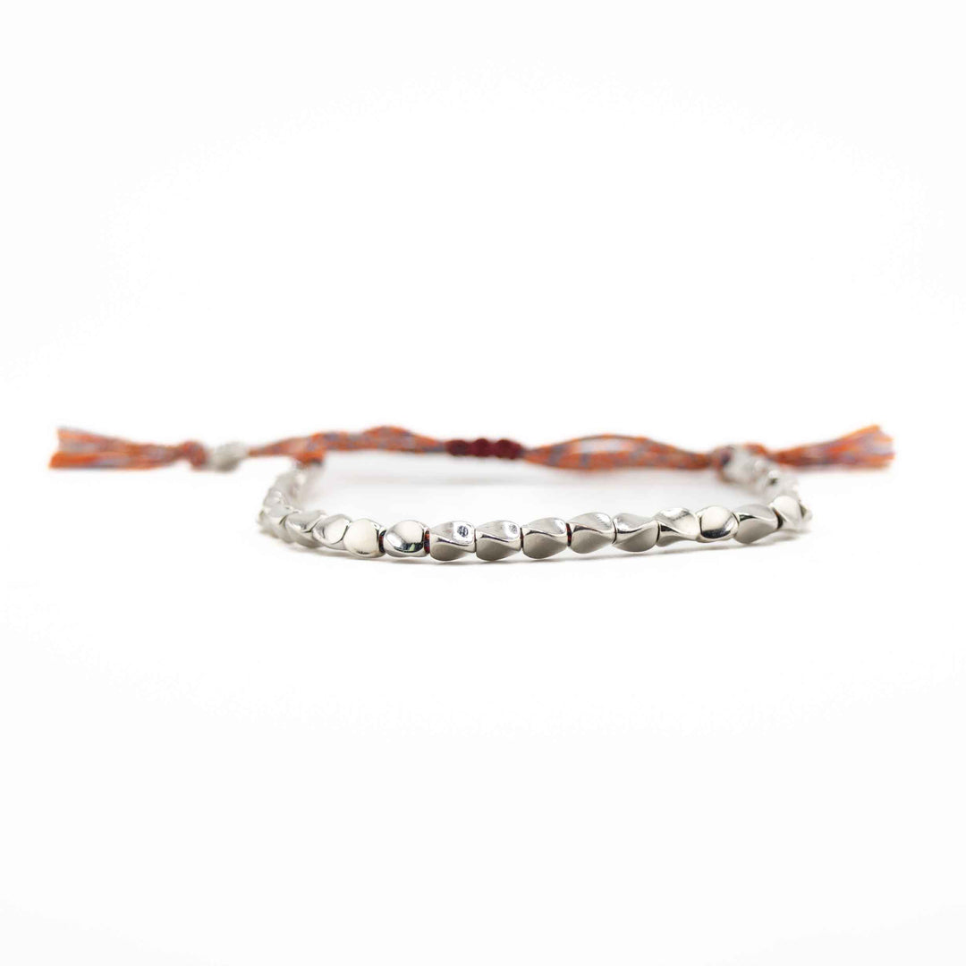 A horizontal, eye-level view of the copper bead bracelet, focusing on the row of shiny, organic-shaped silver beads with the multi-colored cord and tassels blurred in the background.