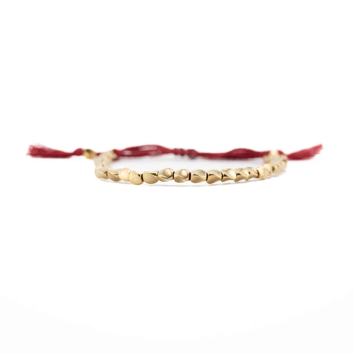 A horizontal, eye-level view of the copper bead bracelet, focusing on the row of shiny, organic-shaped gold beads with the red cord and tassels blurred in the background.