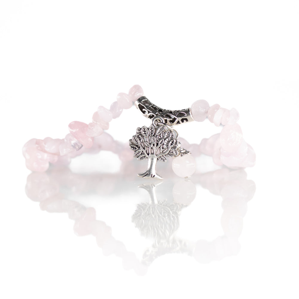 Photograph of a rose quartz chip bead bracelet featuring a detailed silver-toned Tree of Life charm and an ornate silver barrel bead, reflected on a pristine white surface