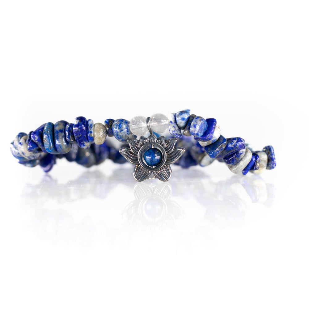 A handcrafted lapis lazuli chip bracelet featuring round clear quartz beads and a silver-toned lotus flower charm with a central blue stone, resting on a white reflective surface