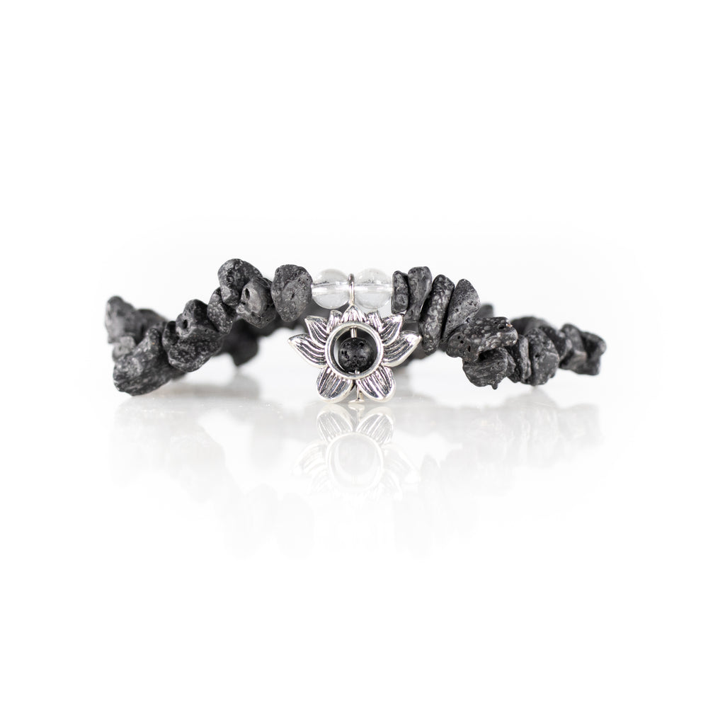 Photograph of a dark grey porous lava stone bracelet with two clear crystal beads and a central silver lotus flower charm on a reflective white surface