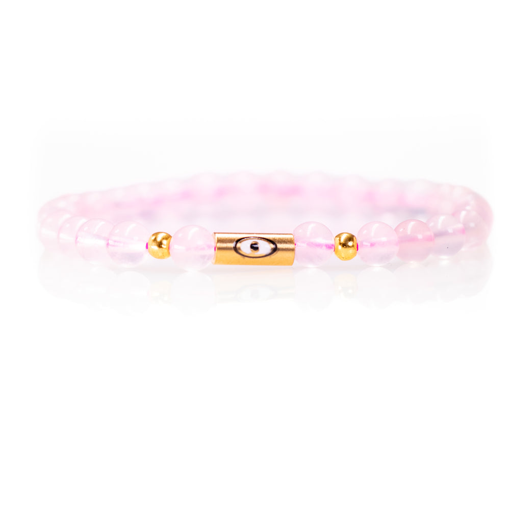 Product photography of a rose quartz bracelet with delicate pink beads, a central gold cylindrical evil eye charm, and two small gold spacer beads on a reflective white surface