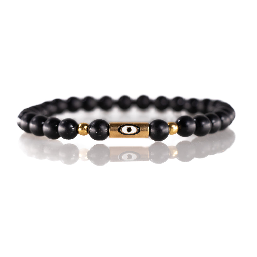 Jewelry a sleek beaded bracelet featuring matte black beads, small gold-tone spacer beads, and a central rectangular gold charm adorned with a white evil eye design on a reflective white surface