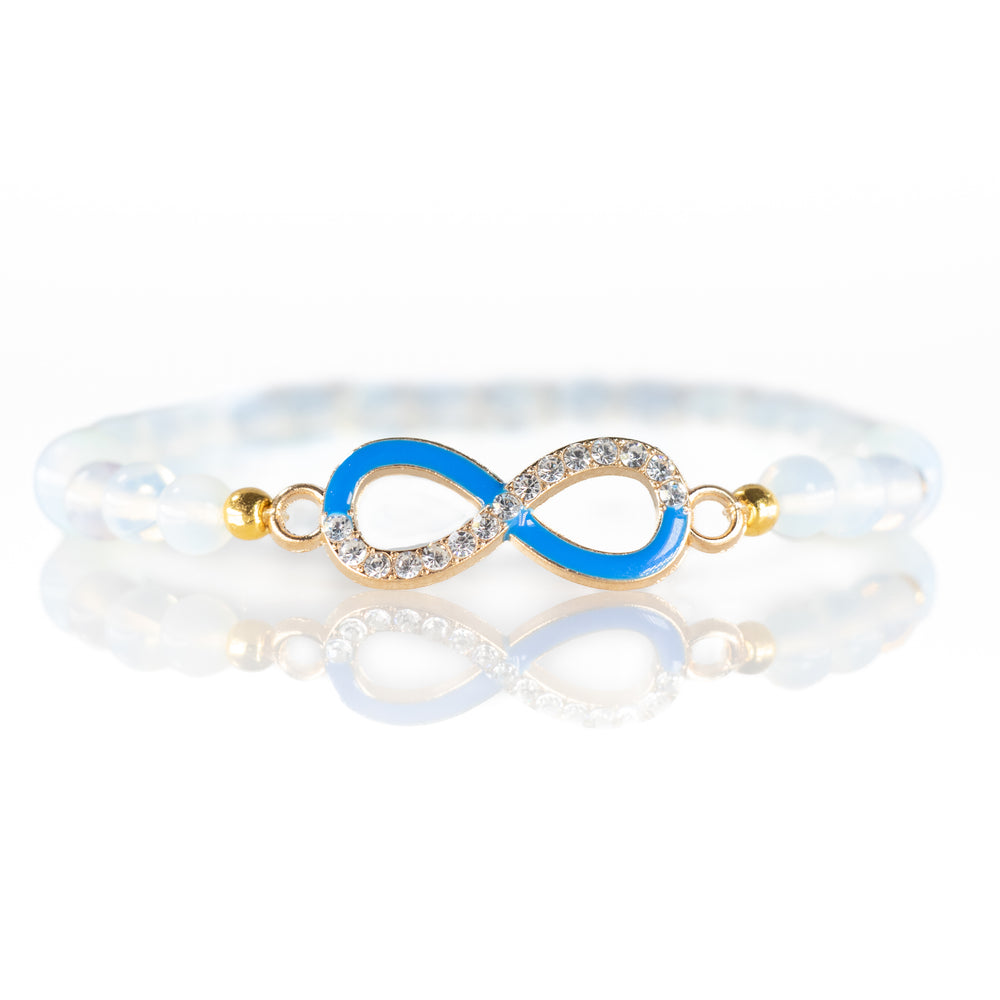 Jewelry: a white opalescent beaded bracelet with a striking gold infinity charm half adorned with brilliant clear rhinestones and half with vivid blue enamel beautifully reflected on a pristine white surface