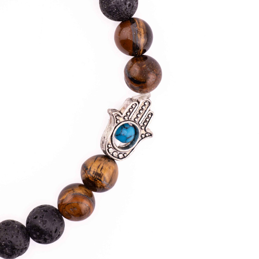 Close-up detail of silver Hamsa hand charm with blue evil eye inlay positioned between golden tiger eye beads on a black lava stone bracelet