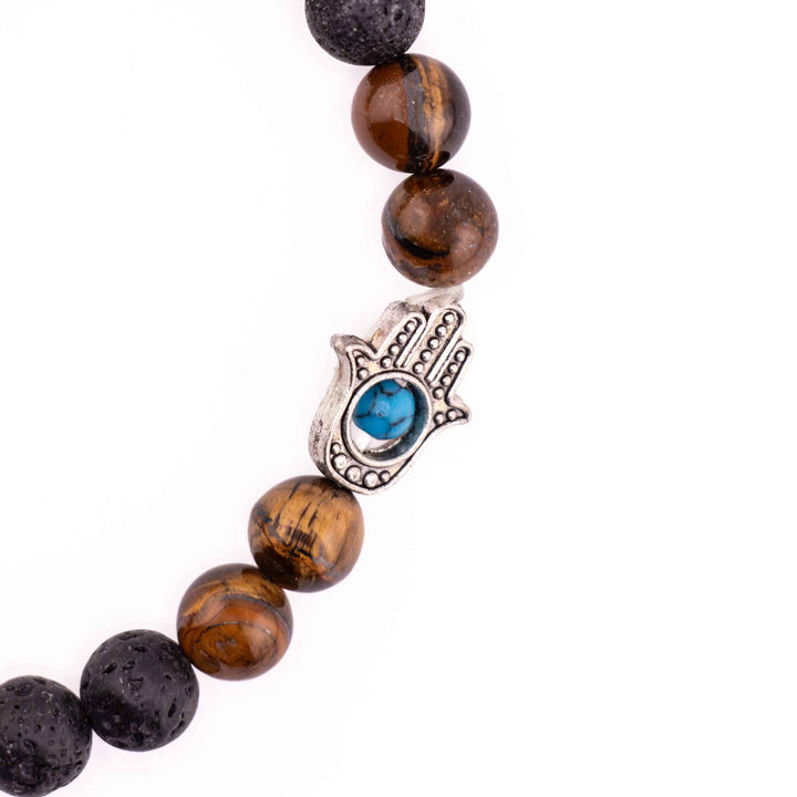 Close-up detail of silver Hamsa hand charm with blue evil eye inlay positioned between golden tiger eye beads on a black lava stone bracelet