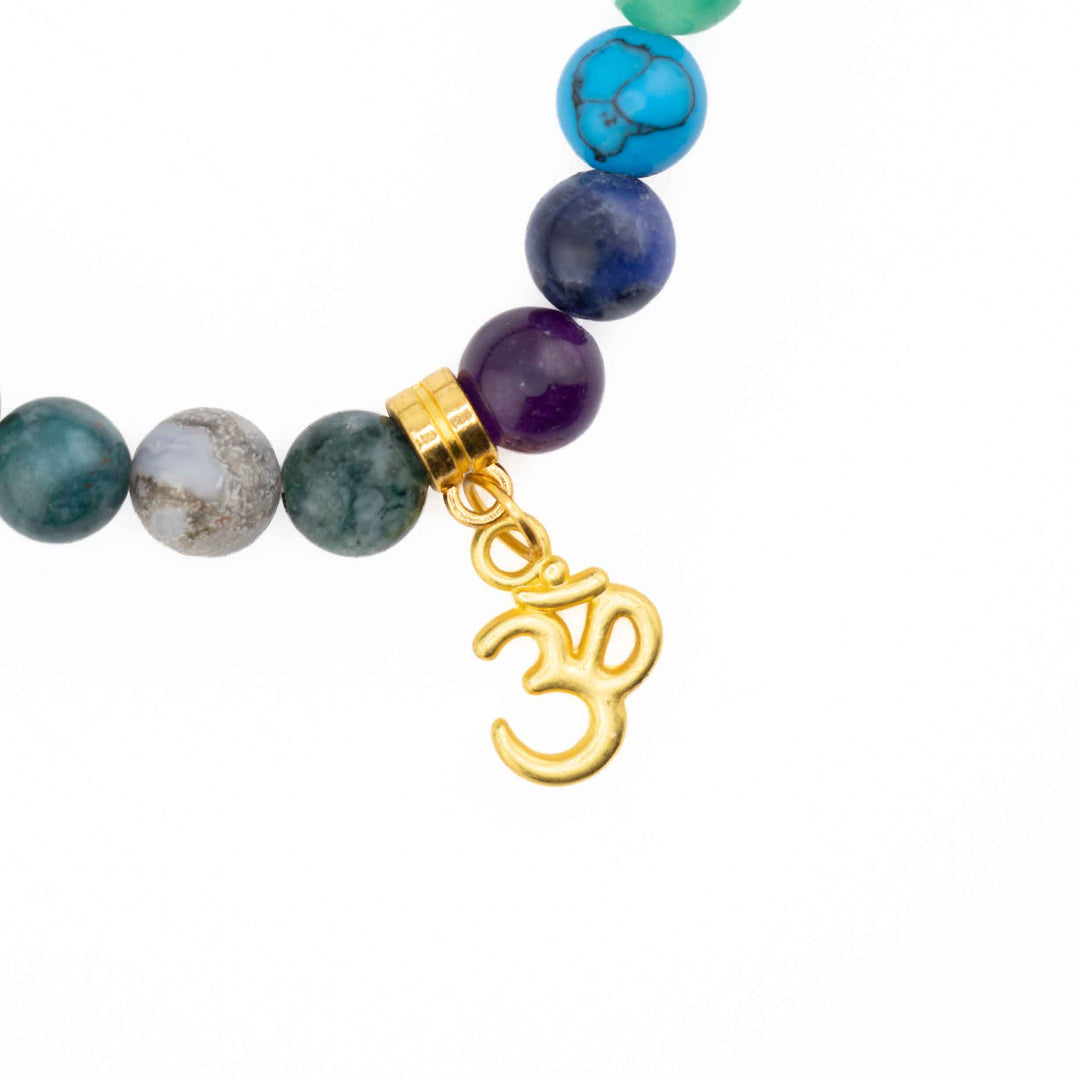Close-up detail of a dangling gold Om charm attached next to purple amethyst, blue lapis lazuli, and turquoise chakra beads on an Indian Agate bracelet