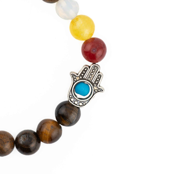 Close-up detail of silver Hamsa hand charm with blue evil eye inlay and multicolored chakra beads on a brown Tiger Eye bracelet