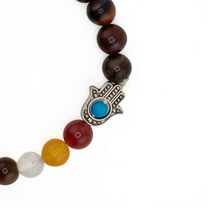 Close-up view of silver Hamsa hand charm with blue evil eye inlay next to vibrant chakra beads on a dark red Tiger Eye bracelet