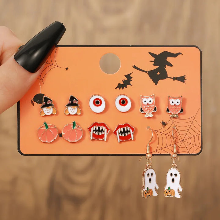 Enamel Halloween themed jewelry set featuring stud earrings of witch heads red eyeballs orange pumpkins vampire lips and owls along with ghost dangle earrings displayed on an orange card adorned with a flying witch silhouette bats and spiderwebs