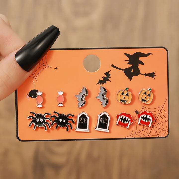 A Halloween stud earring set on an orange display card held by a hand with a black coffin nail, featuring black witches, orange jack-o'-lanterns, and vampire fangs